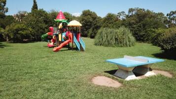 A playground with slides and climbing structures, surrounded by grass and trees.