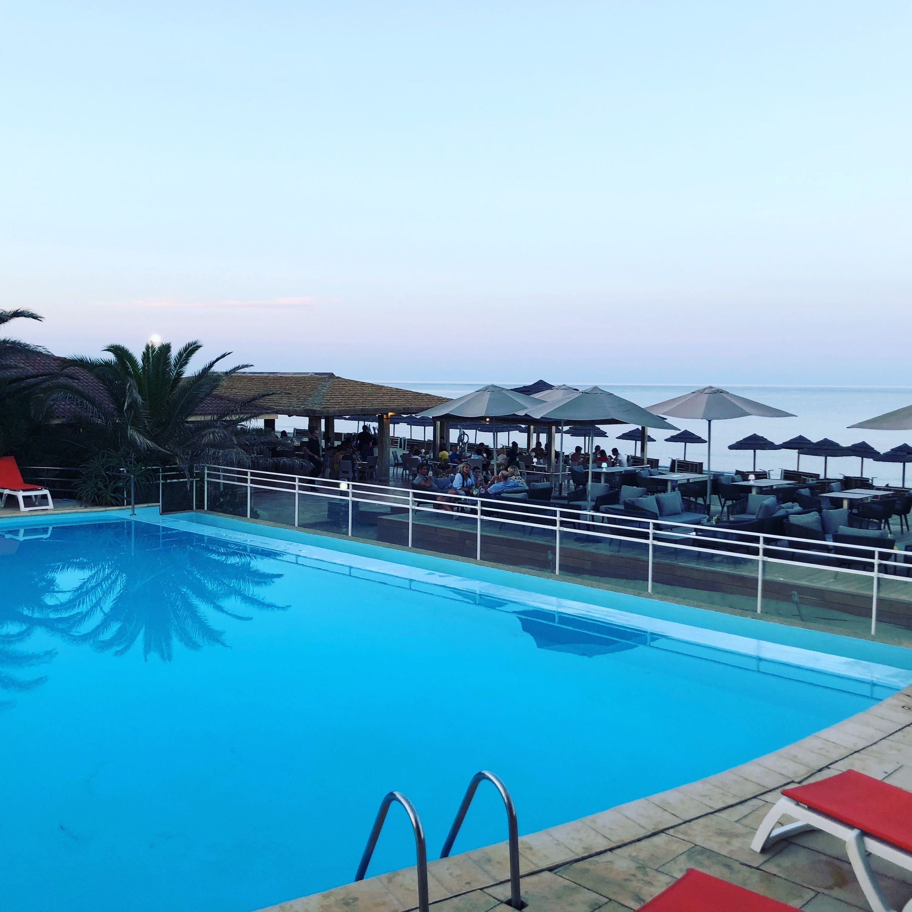 A poolside area with lounge chairs and umbrellas, a clear sky, and a view of the sea.