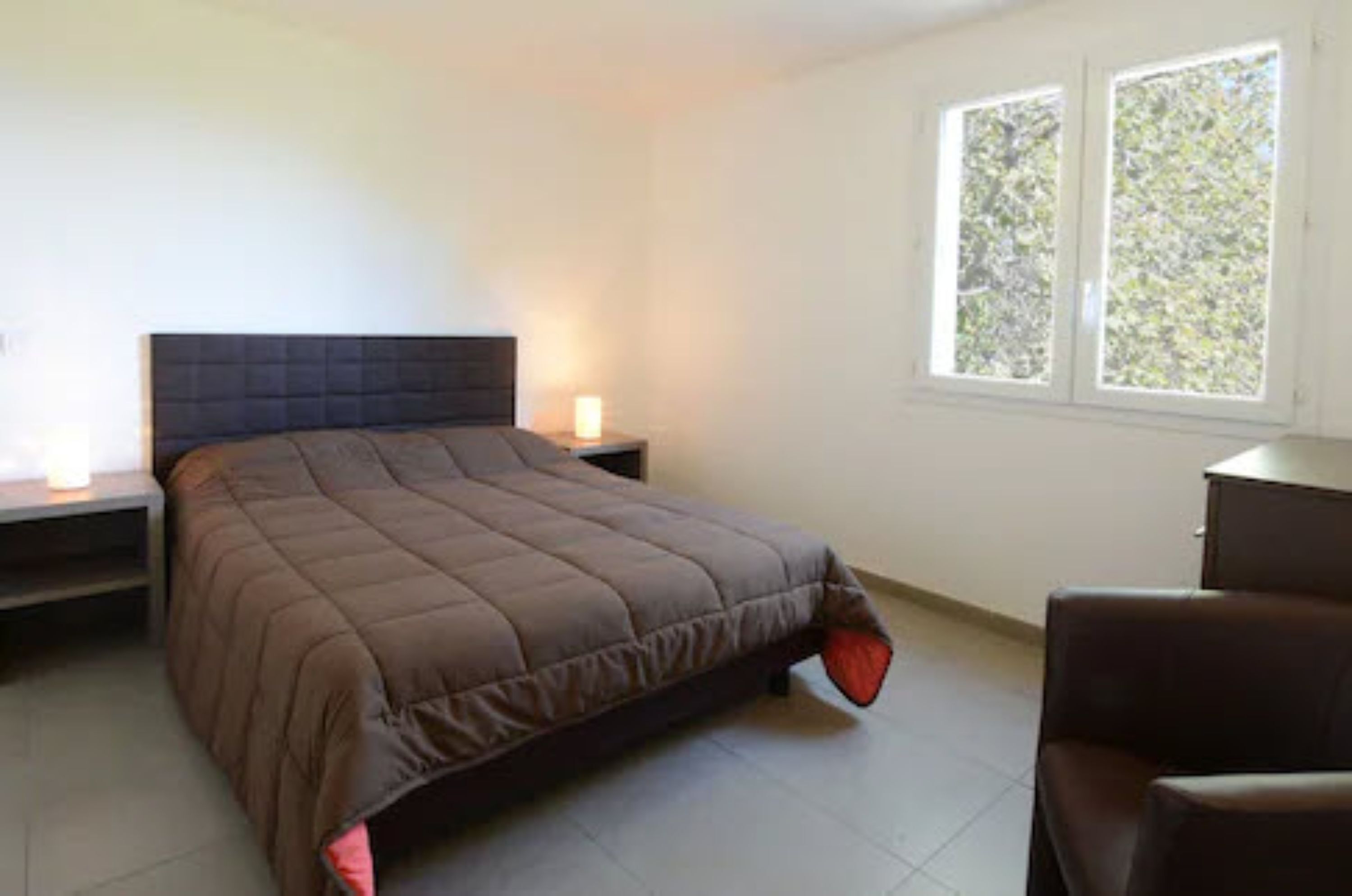 Une chambre à coucher avec un lit, des tables de chevet, une chaise et une fenêtre donnant sur des arbres.