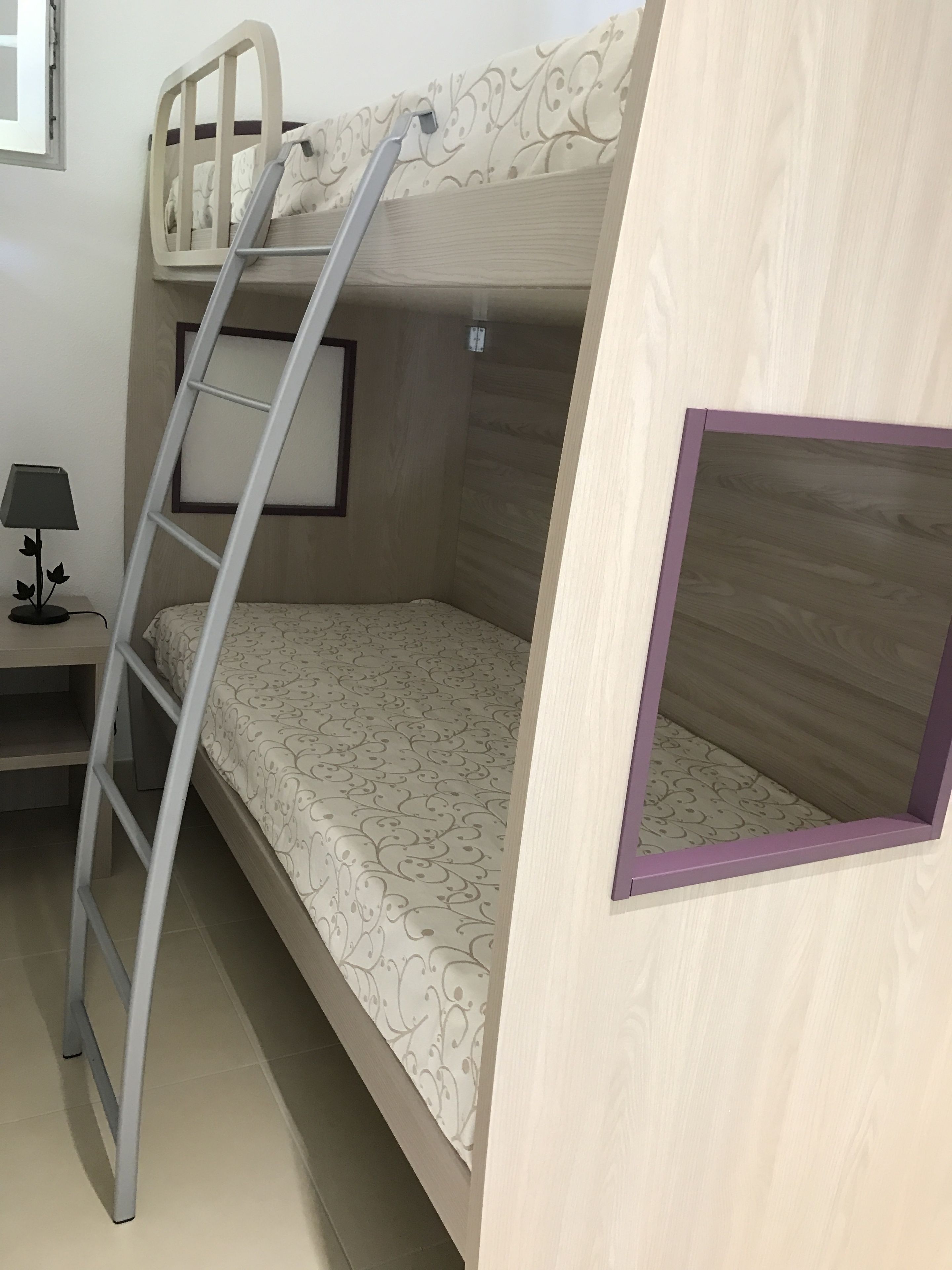 Un lit superposé avec une échelle, une petite table avec une lampe et une fenêtre.