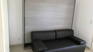 A modern room with a black leather sofa against a light-colored wall, a large window, and a tiled floor.