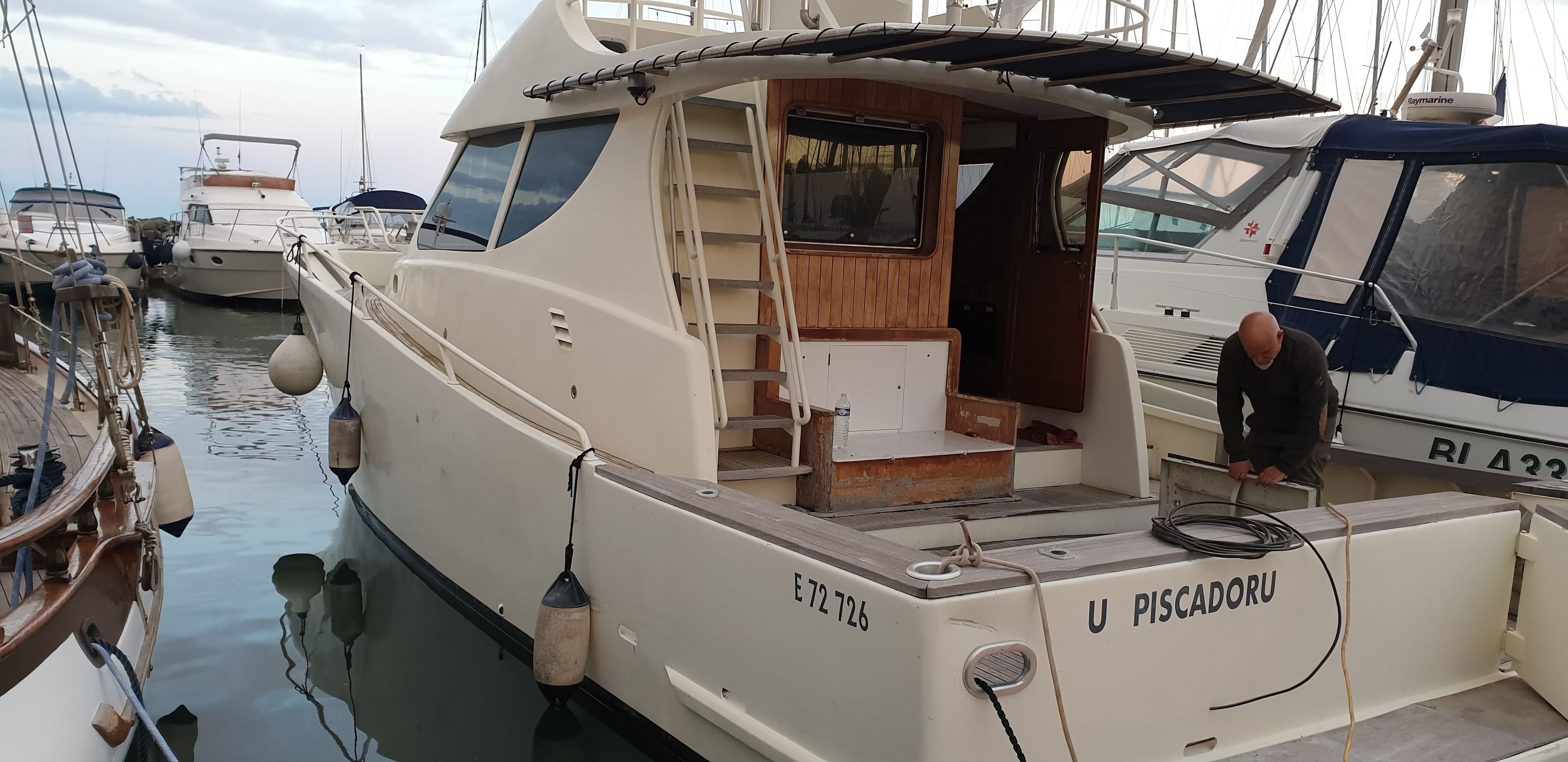 Een boot genaamd 'U PISCADORU' afgemeerd in een jachthaven, met andere boten, een ladder naar het dek en een hut met een raam.