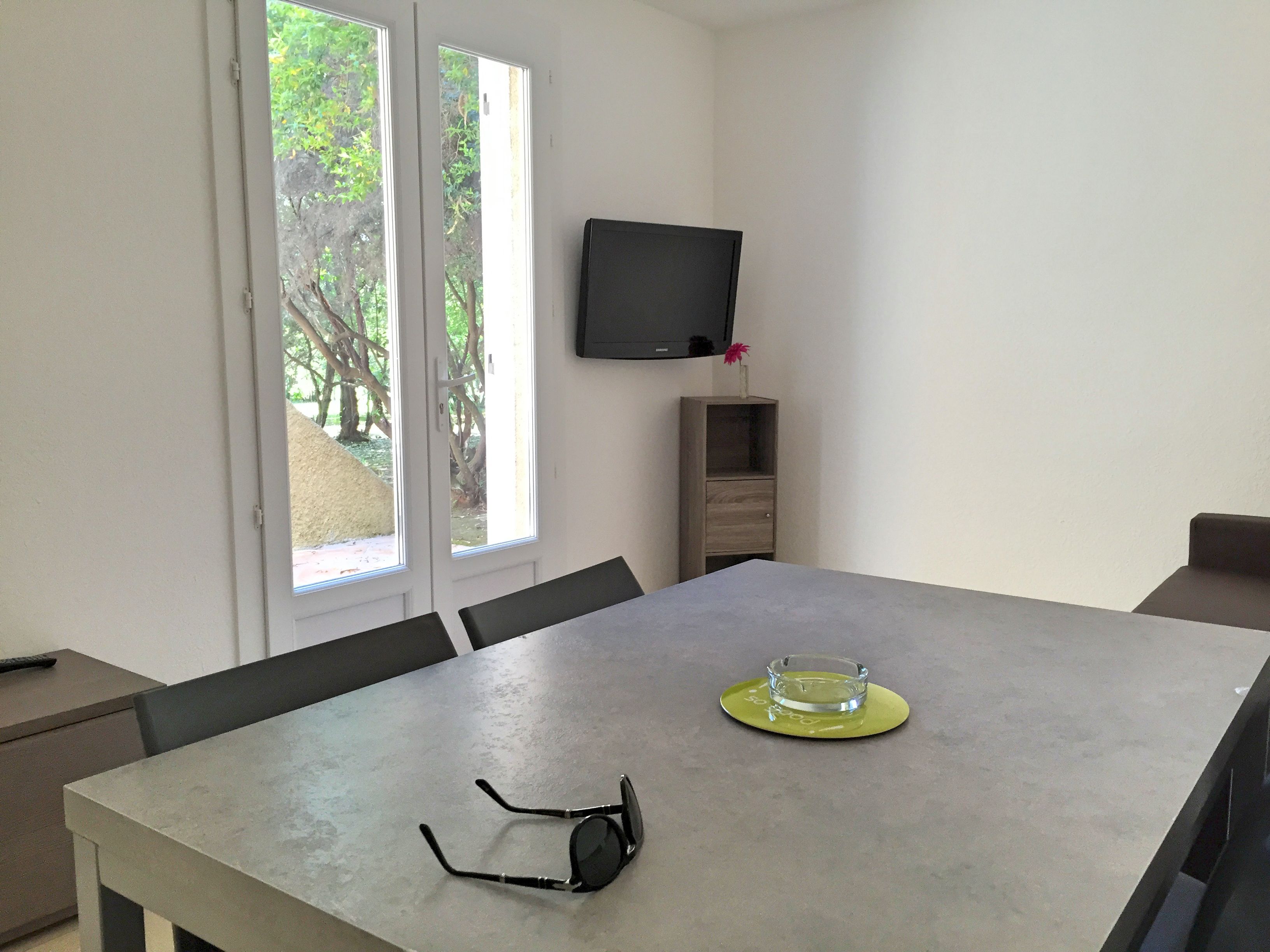 Un coin repas comprenant une table, des chaises, un téléviseur fixé au mur et une fenêtre donnant sur la forêt.