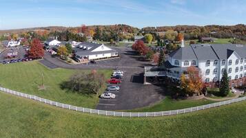An aerial view of a large building complex with multiple parking areas, surrounded by green fields and trees with autumn foliage.