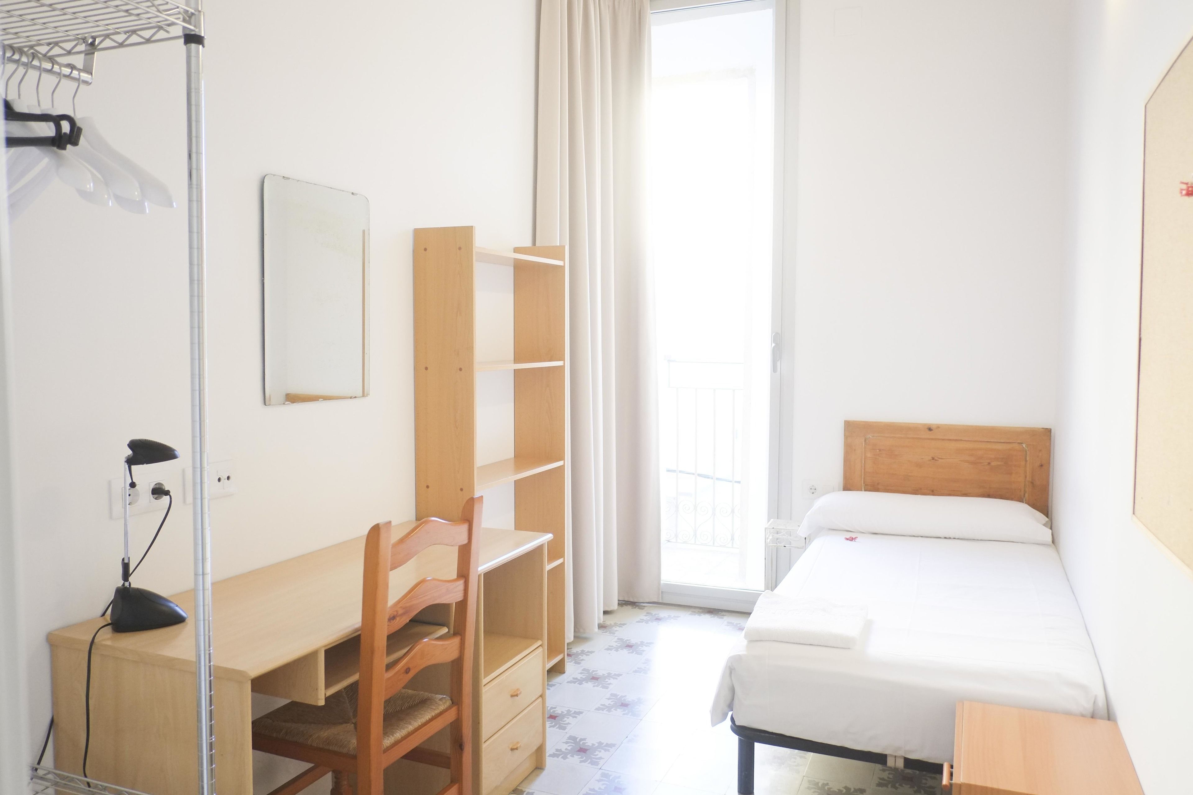A bedroom with a wooden desk, chair, and shelves, a bed with a wooden headboard, and a window with curtains.
