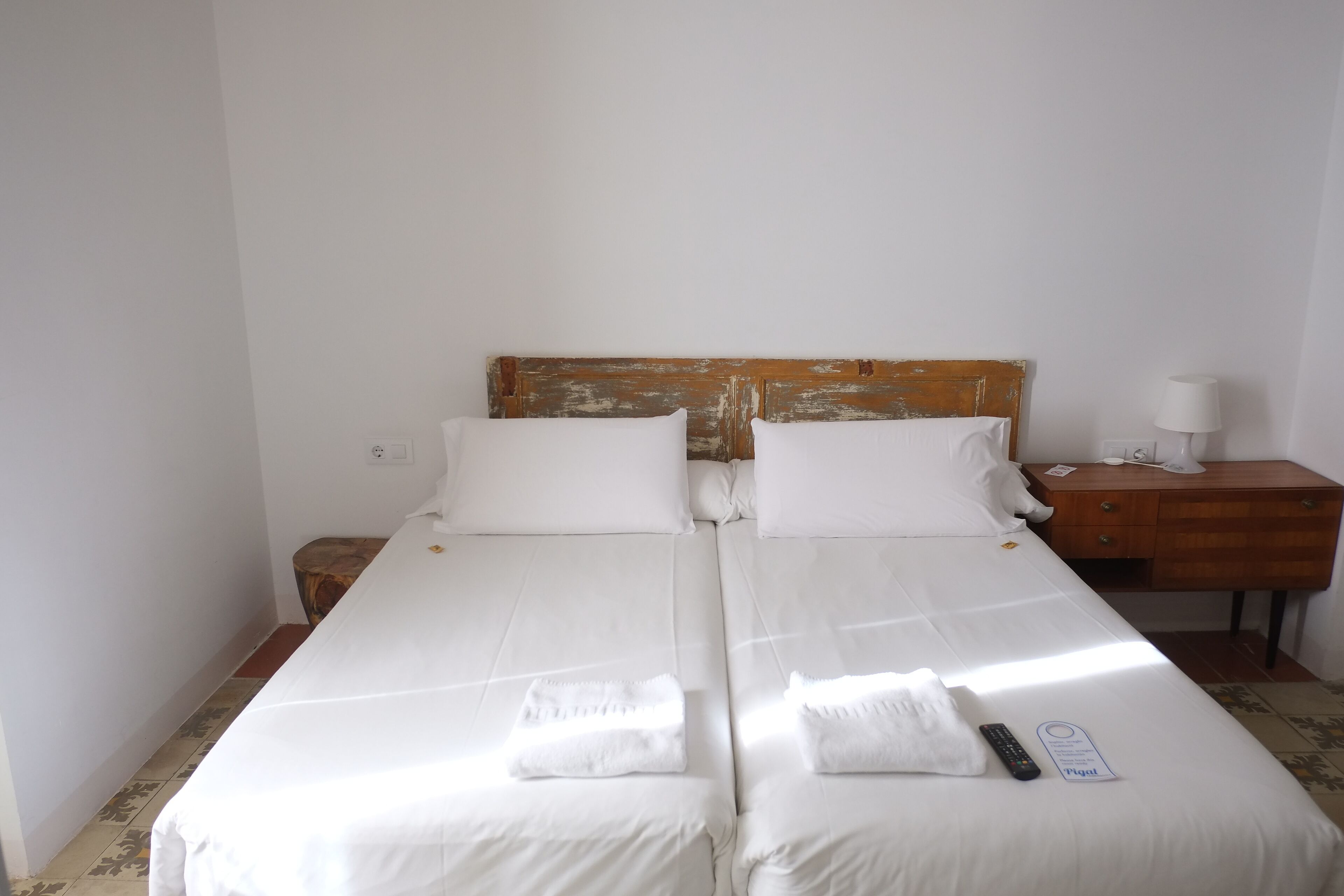 A double bed with white linens, a wooden headboard, a bedside table with a lamp, and a remote control on the bed.