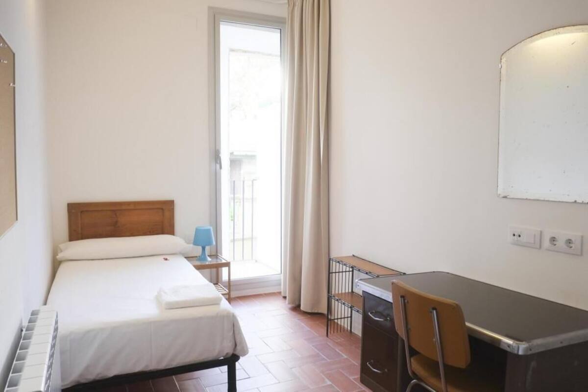Un dormitorio con cama, escritorio, silla, lámpara y vistas por la ventana.