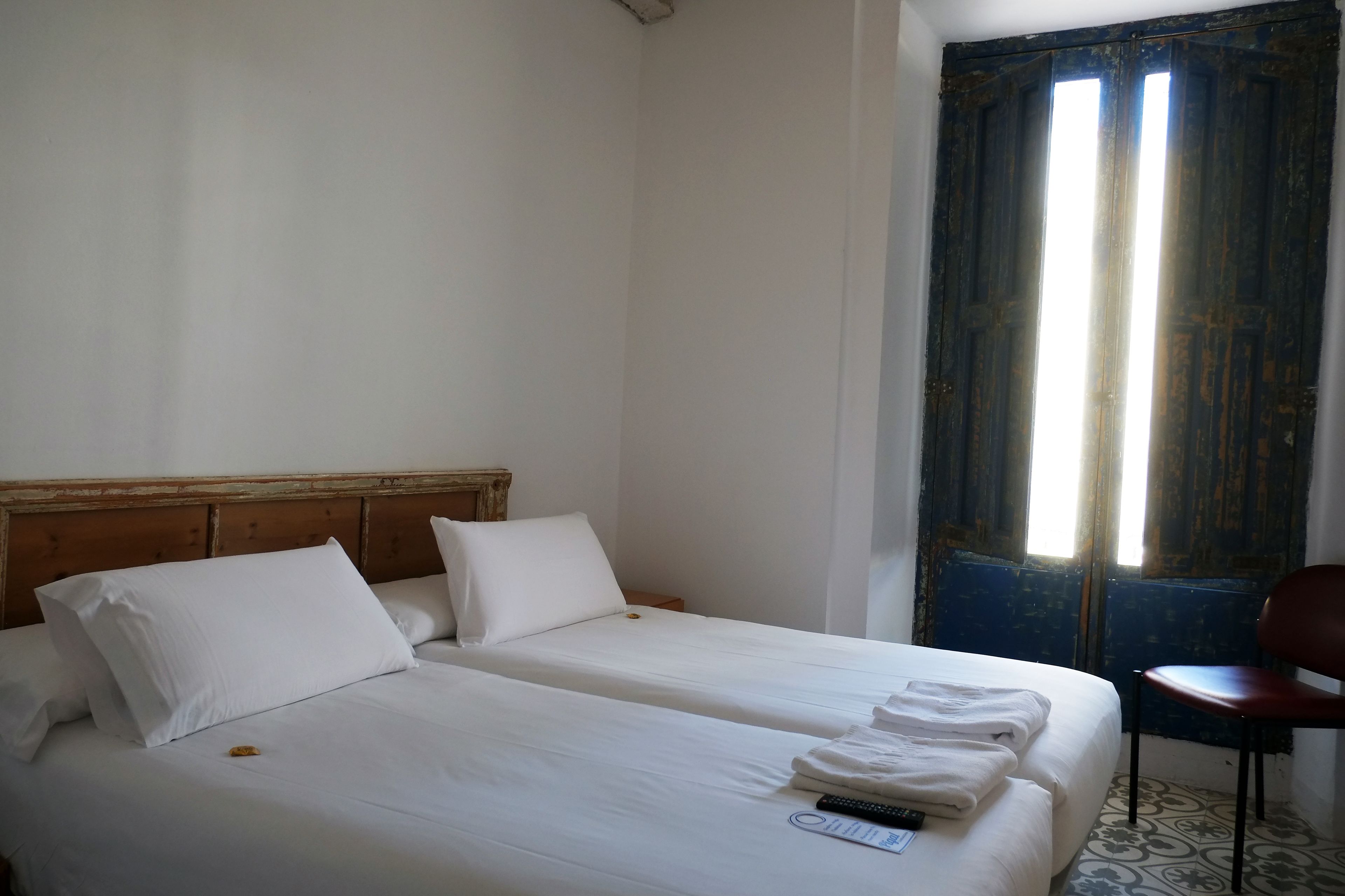 Habitación de hotel con dos camas, cabecero de madera, una silla y un ventanal con cortinas.