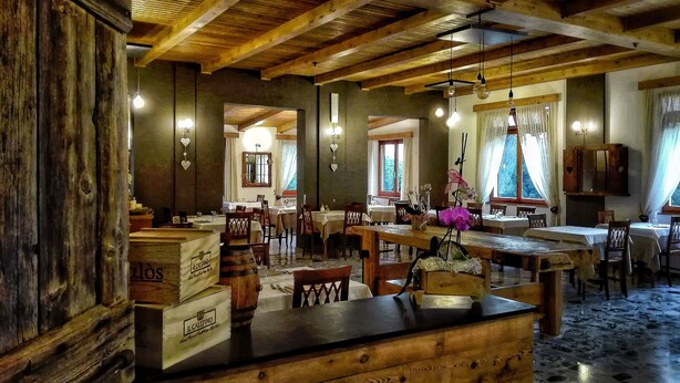 Un comedor rústico con mesas y sillas de madera, una barra con cajas etiquetadas “IL CALPINO”, y vista del salón comedor a través de un vano.