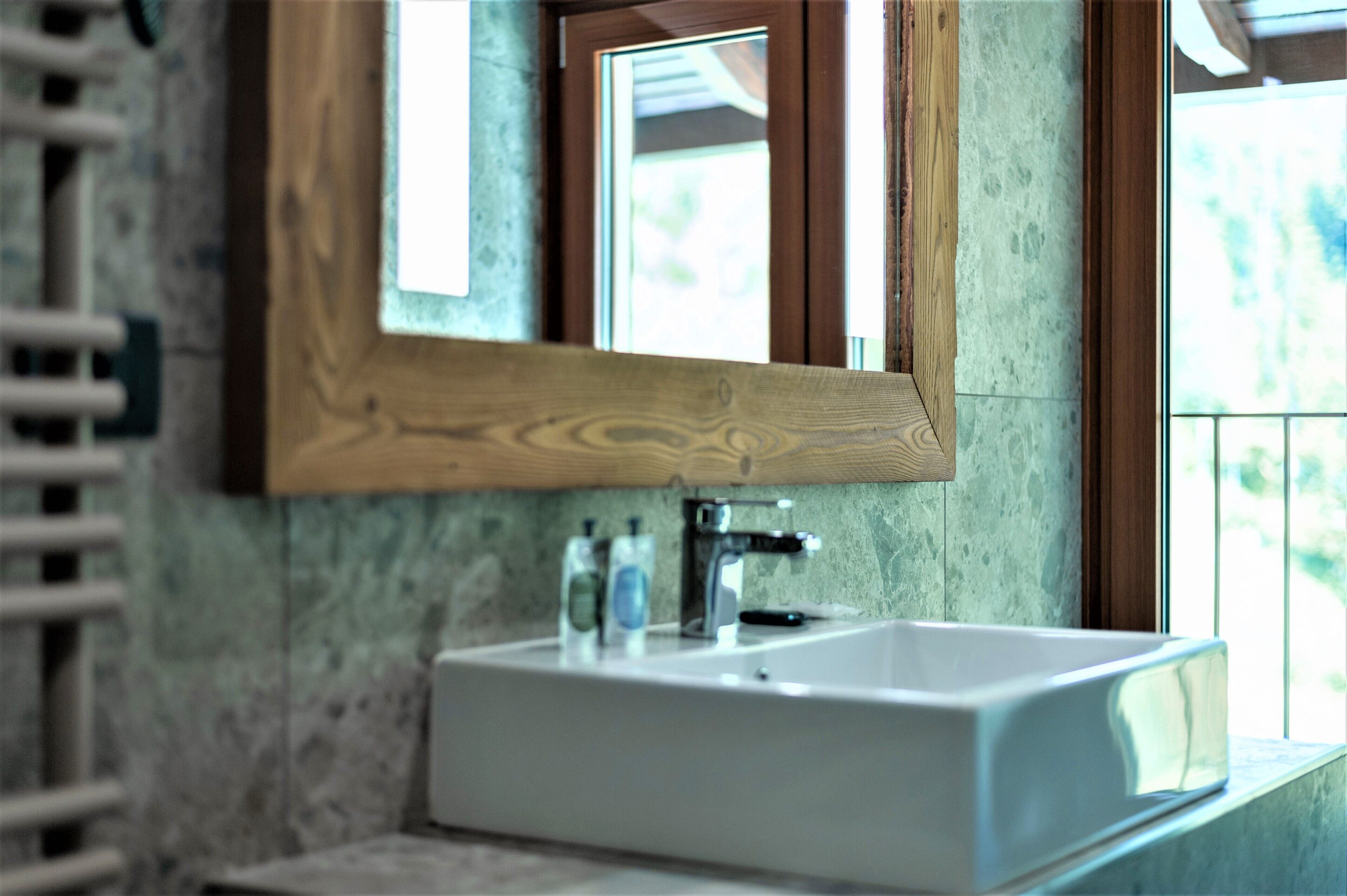 A bathroom with a white sink, wooden-framed mirror, and a window with a view of greenery.