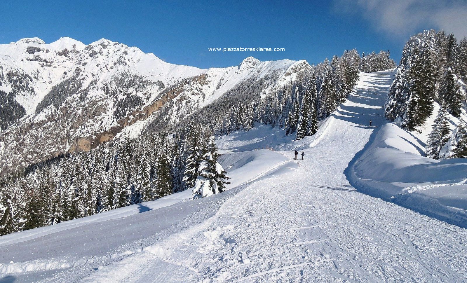 A snowy mountain landscape with a ski slope, trees, and a clear sky.