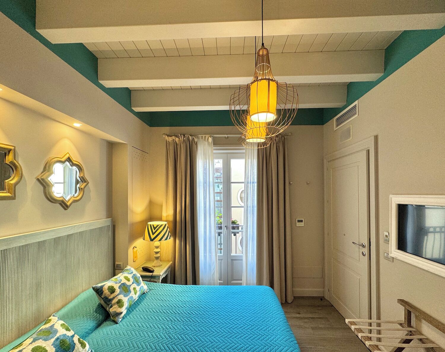 A hotel room with a bed, a TV, a lamp, and a door leading to a balcony.