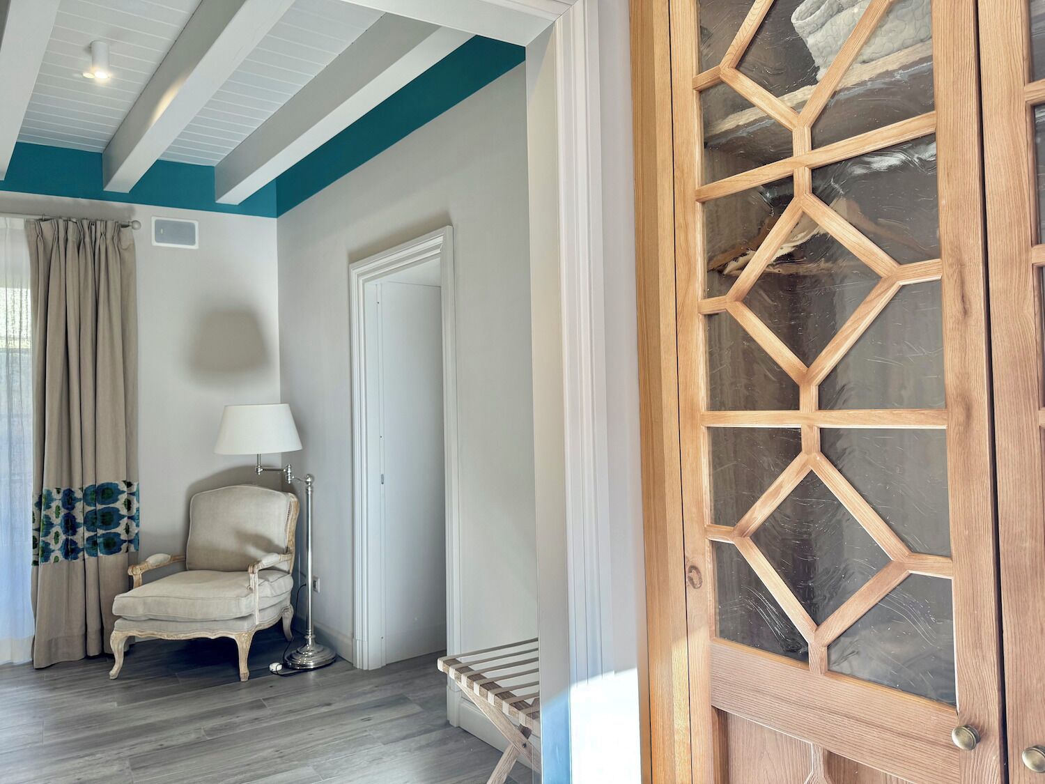 Una habitación con puerta de madera, una silla, una lámpara y un ventanal con cortinas.
