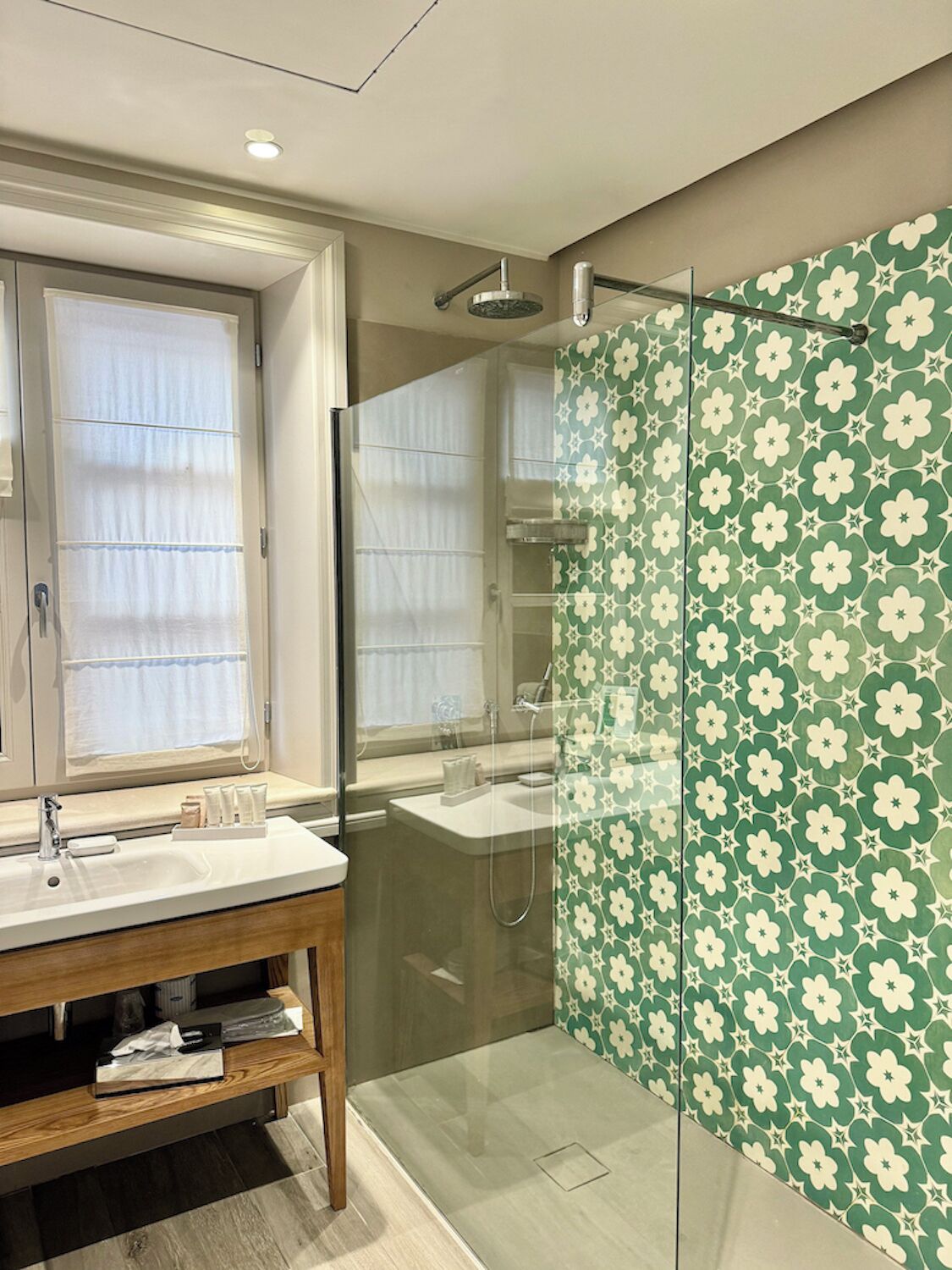 A modern bathroom with a glass-enclosed shower, a vanity with a mirror, and a window with sheer curtains.