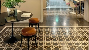 A modern interior with a geometric patterned floor, a round table with two stools, a potted plant, and a decorative wall piece. - Hotel San Michele (Trapani)