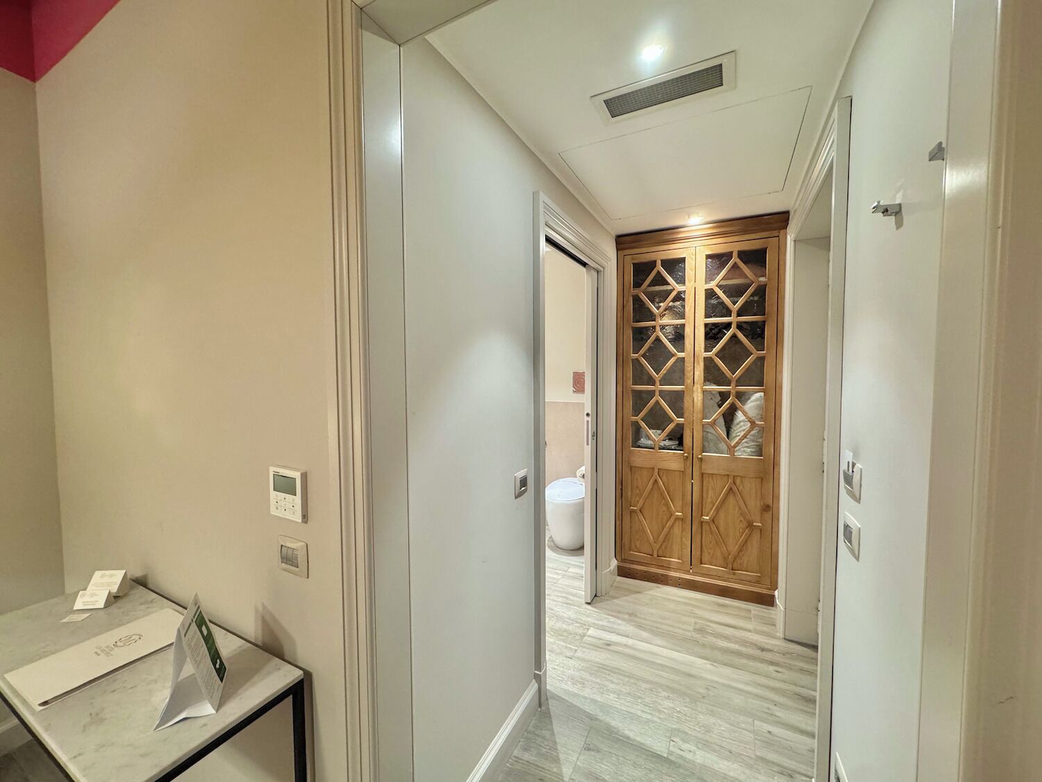 Un pasillo con suelo de mármol, una puerta de madera con un diseño geométrico y una pared blanca con un termostato y un pequeño letrero.