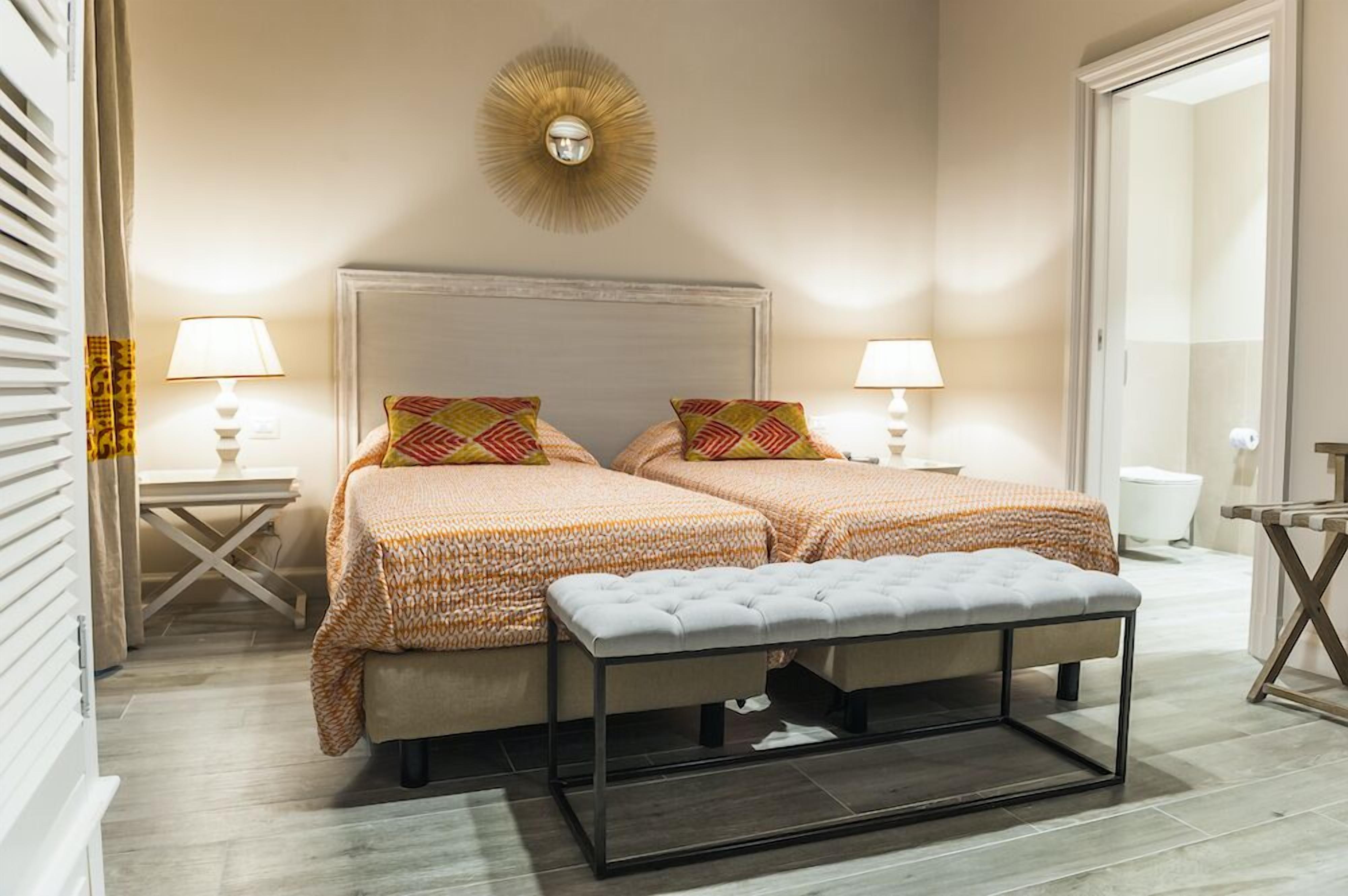 Habitación de hotel con dos camas, un banco, una lámpara de pared y una pequeña mesa con lámpara.