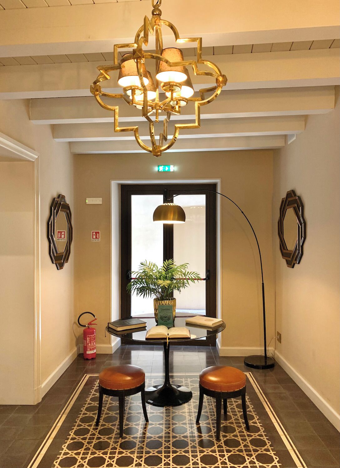 A hallway with a geometric patterned floor, a central table with a potted plant, two stools, a tall floor lamp, and a decorative chandelier.