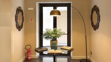A hallway with a geometric patterned floor, a central table with a potted plant, two stools, a tall floor lamp, and a decorative chandelier.