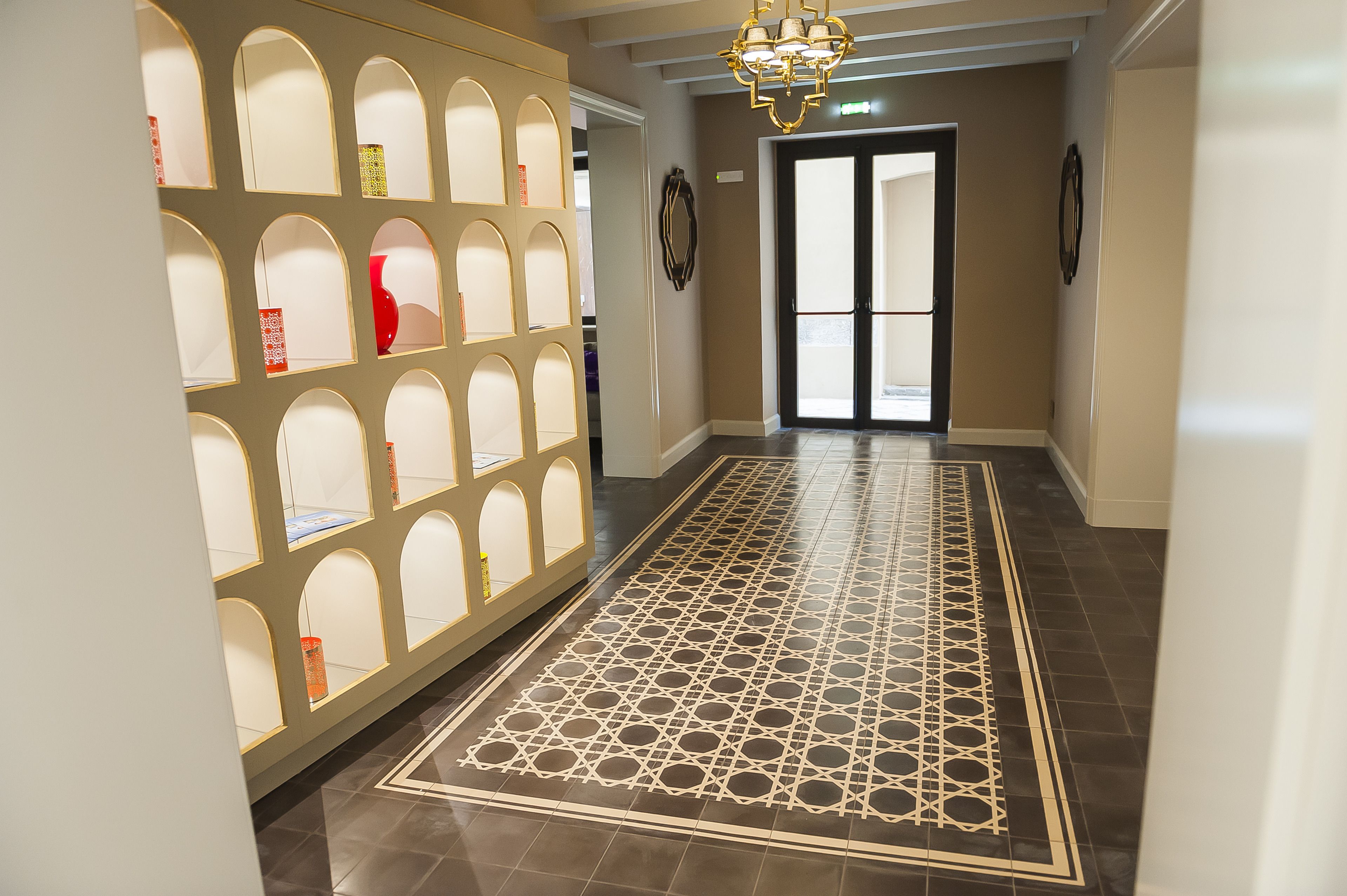 A hallway with a patterned floor, a decorative wall with multiple arched niches, and a chandelier.