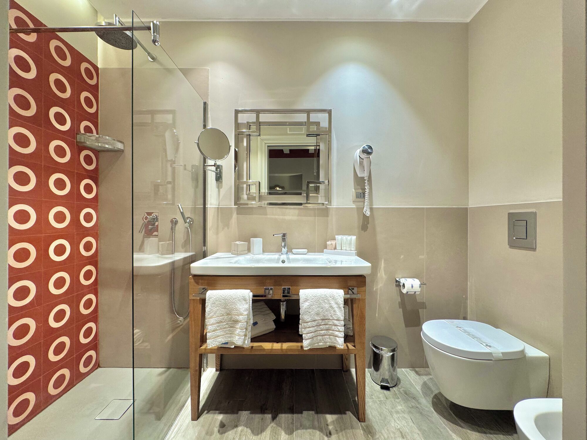 Un baño moderno con cabina de ducha de vidrio, tocador de madera con dos toallas, inodoro blanco y espejo sobre el lavabo.