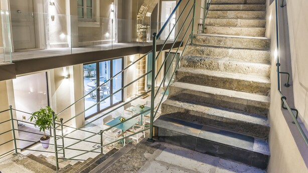 A grand staircase with a glass balustrade, leading to a lower level with a dining area and a potted plant. - Hotel San Michele (Trapani)