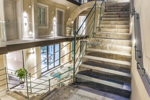 A grand staircase with a glass balustrade, leading to a lower level with a dining area and a potted plant. - Hotel San Michele (Trapani)