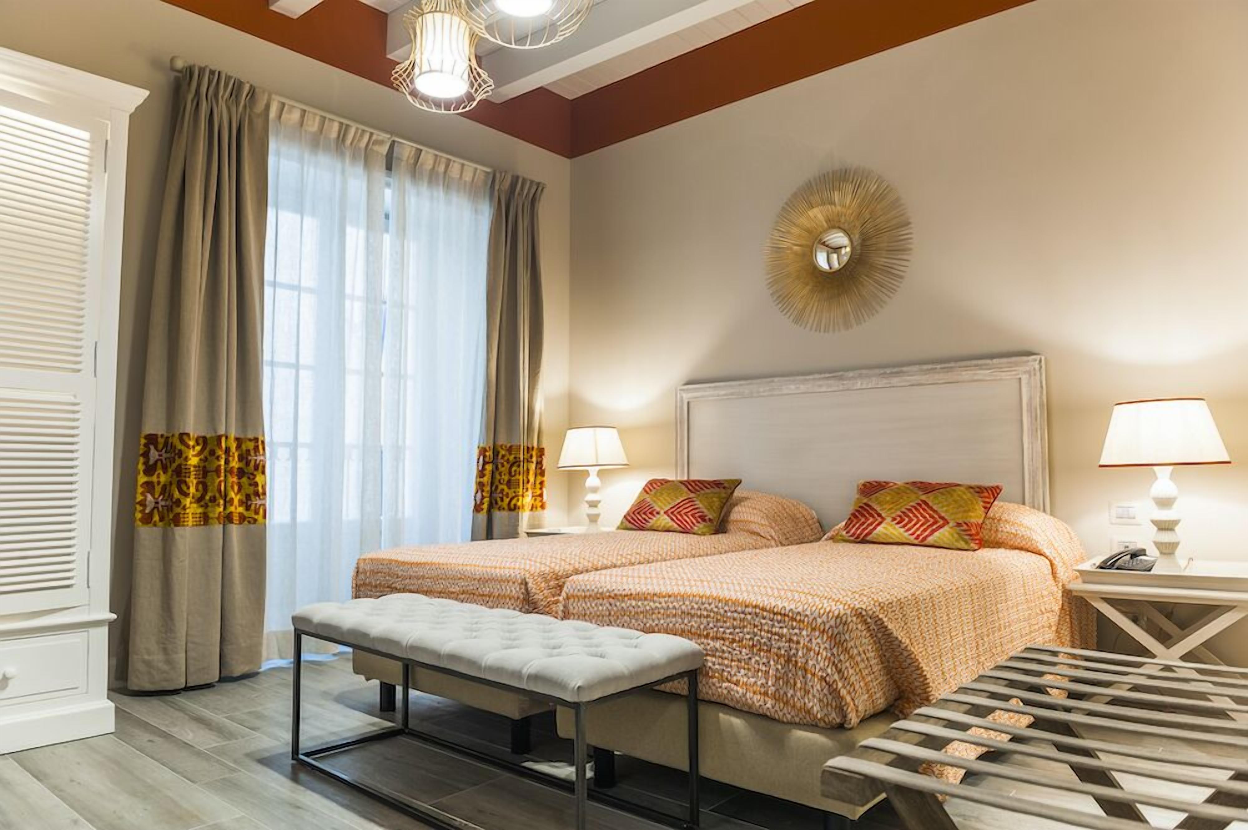 A hotel room with two beds, a bench, a bedside table with a lamp, and a decorative wall piece.