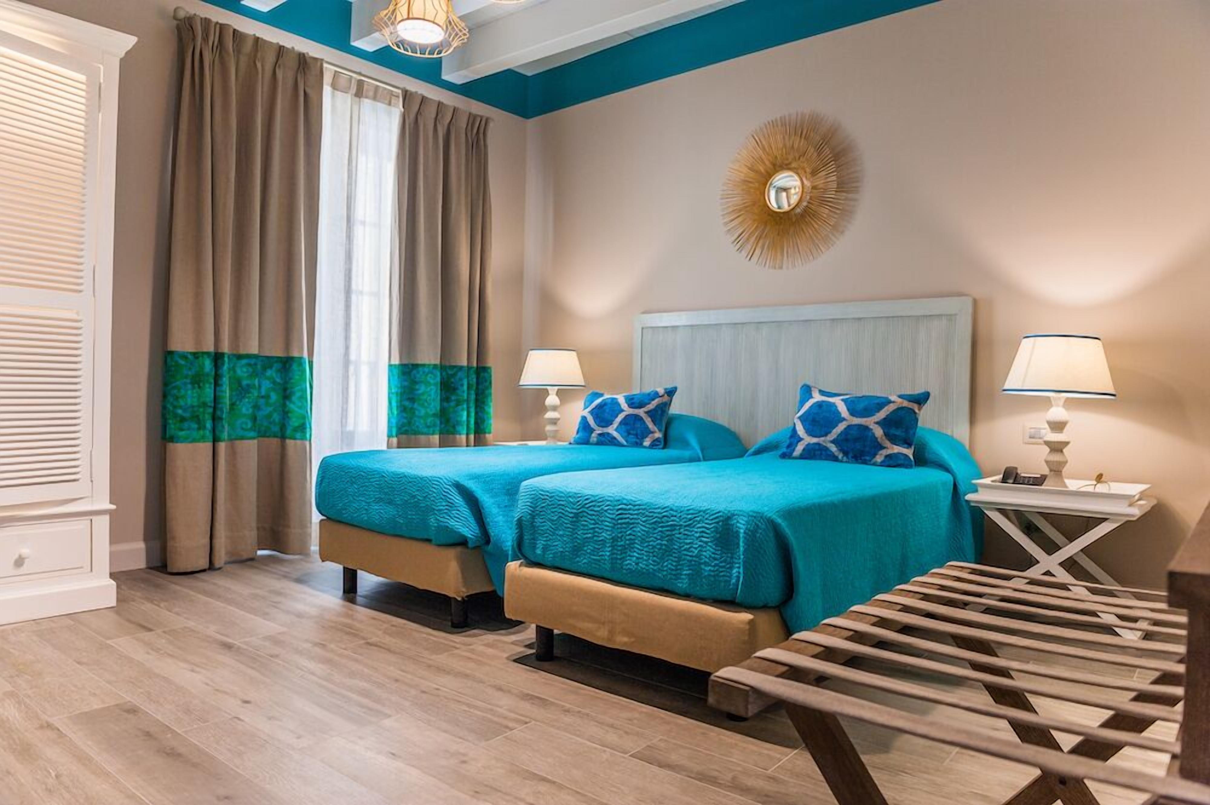 A hotel room with two beds, a wooden bench, a ceiling fan, and a wall-mounted mirror.