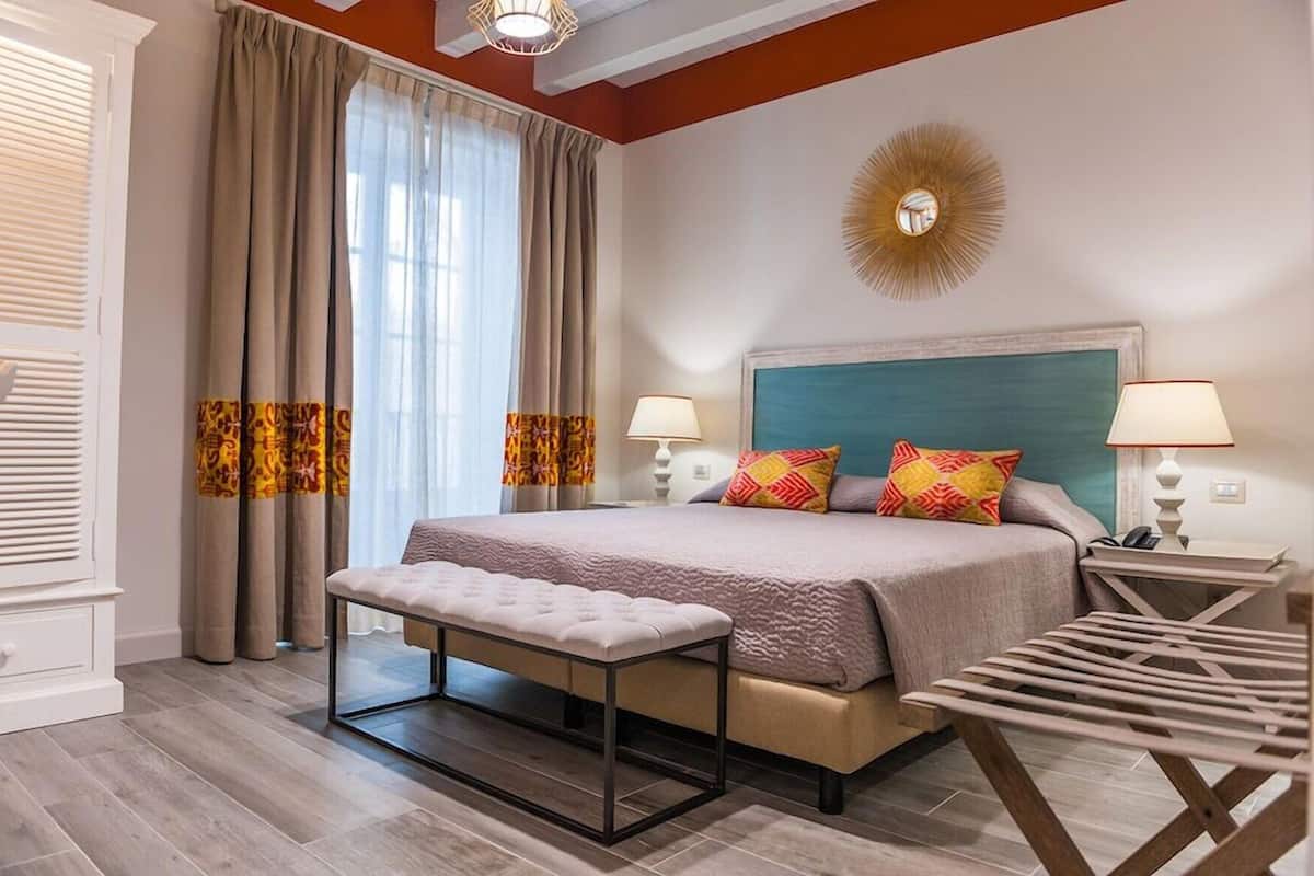 A bedroom with a bed, bench, two bedside tables, a nightstand, a wall-mounted light, and a window with curtains.