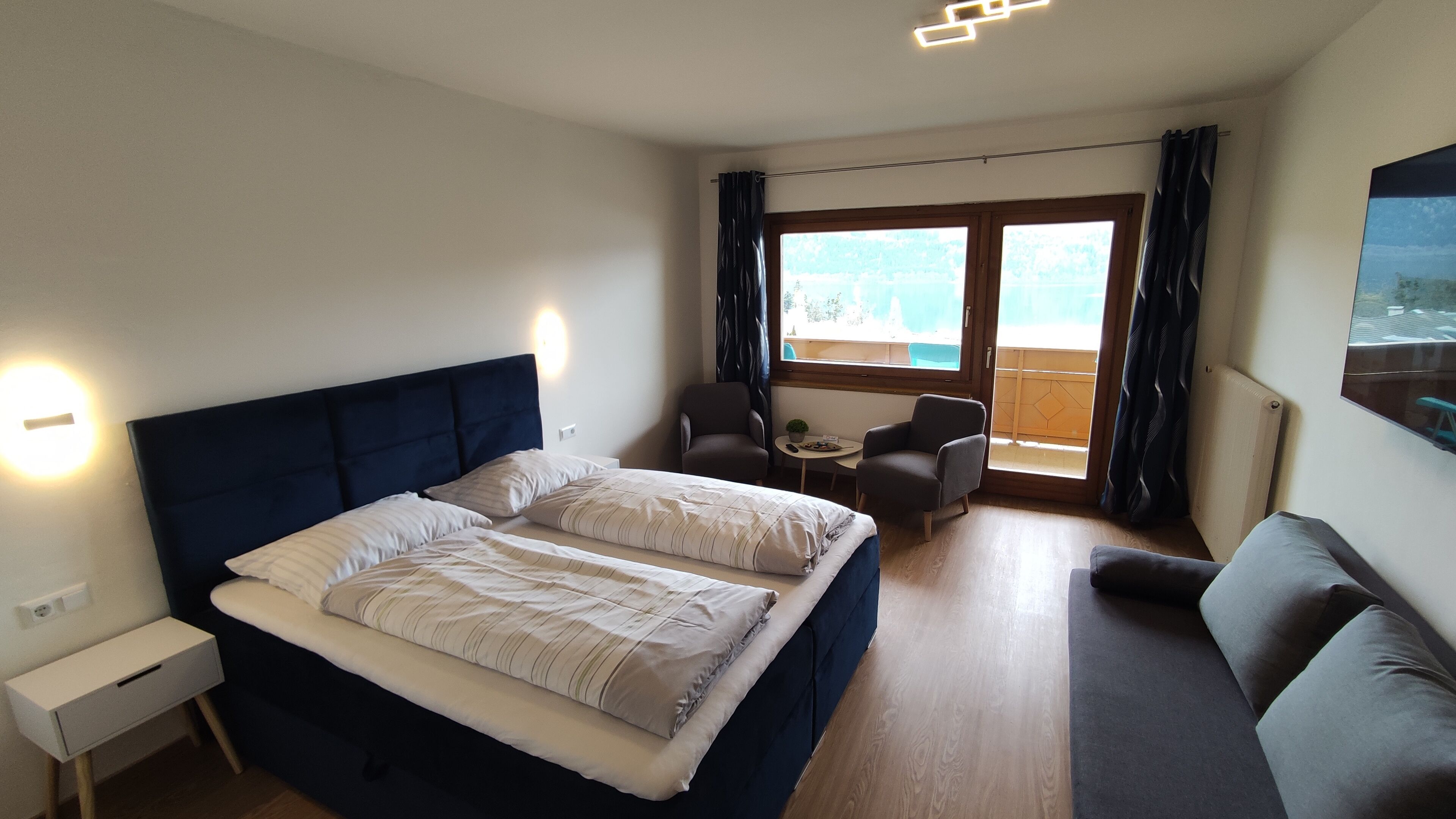 A hotel room with a large bed, a small table with chairs, a sofa, and a balcony with curtains.