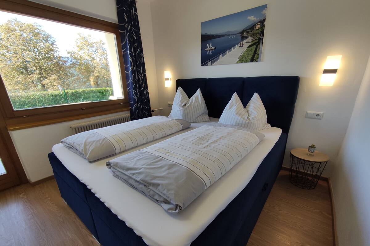 A double bed with a dark headboard, white bedding, and a view of a lake through a window.