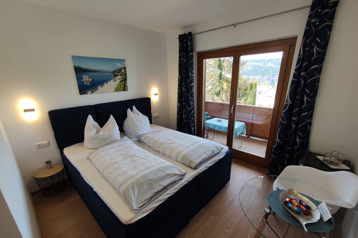 A bedroom with a bed, a painting, a table with glasses, and a view of a balcony.