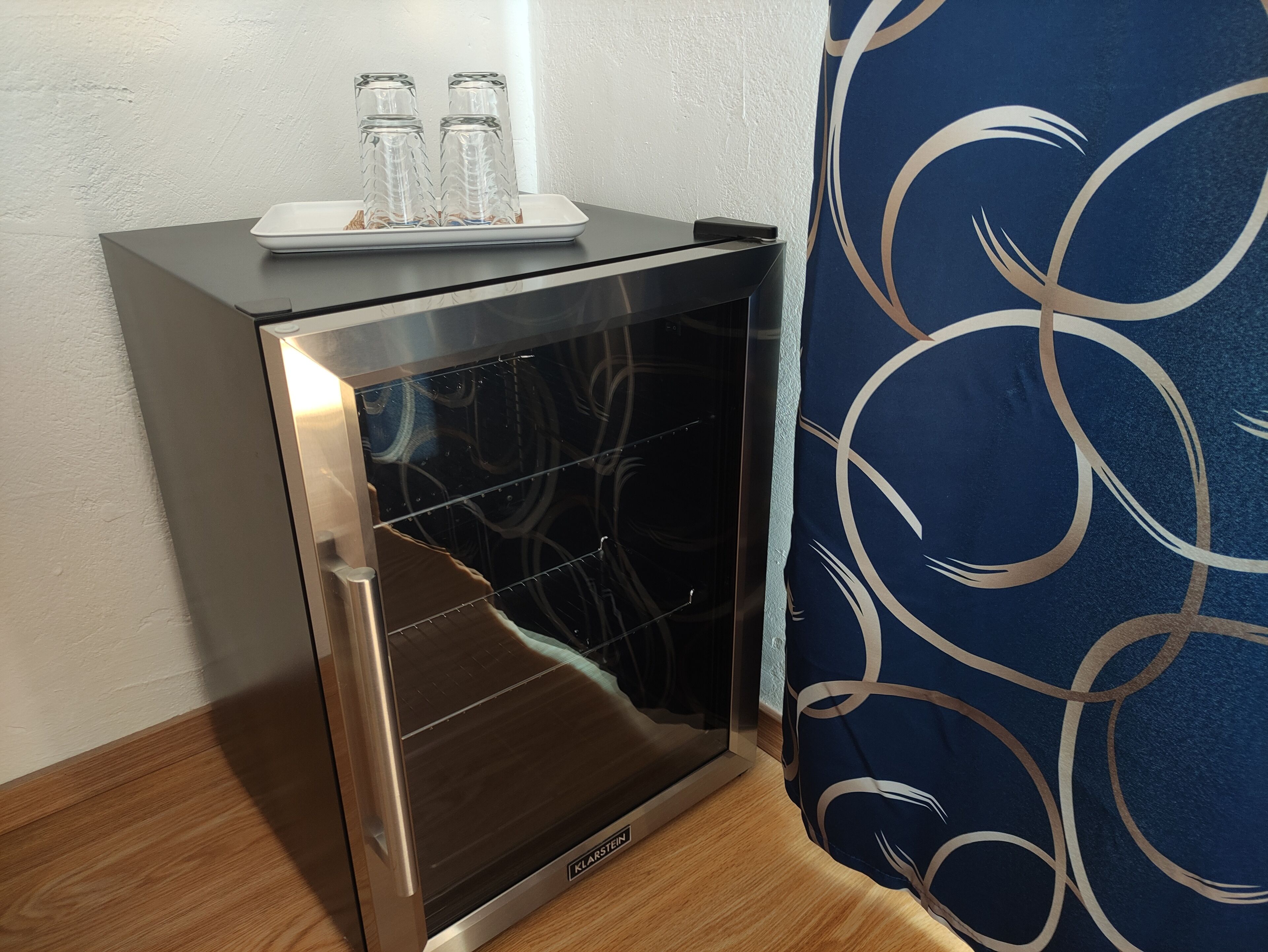 Un mini-réfrigérateur compact en acier inoxydable, doté d’une porte en verre, est placé sur un plancher en bois, à côté d’un rideau bleu orné d’un motif abstrait blanc.