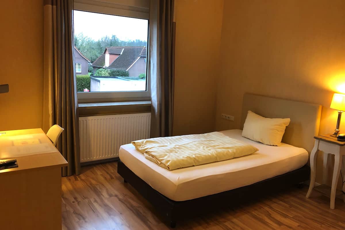 A hotel room with a bed, a desk, a window with curtains, and a bedside lamp.