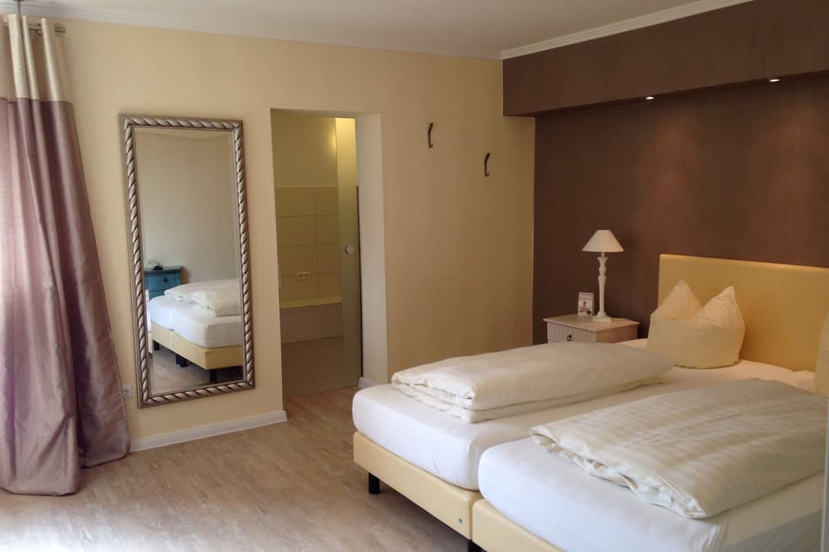 A hotel room with two beds, a large mirror, a bedside table with a lamp, and a bathroom door.