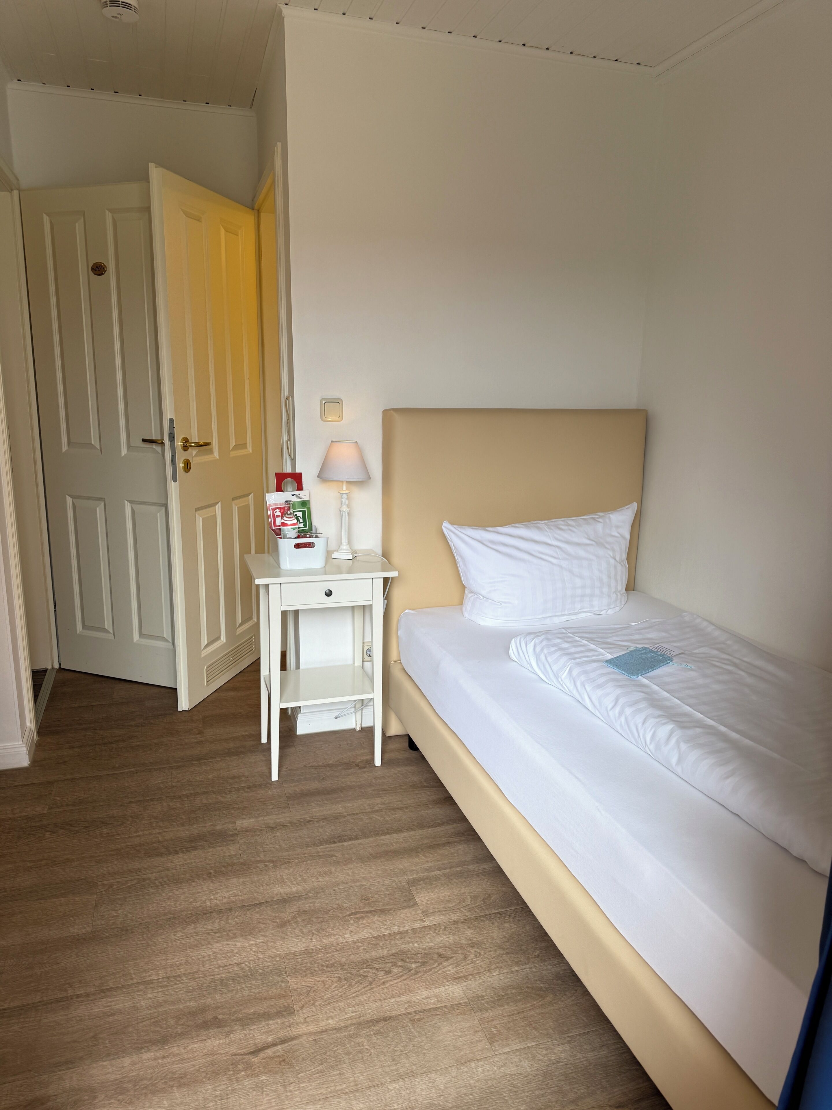 Habitación de hotel con cama, mesita de noche, lámpara y una puerta que da a otra habitación.
