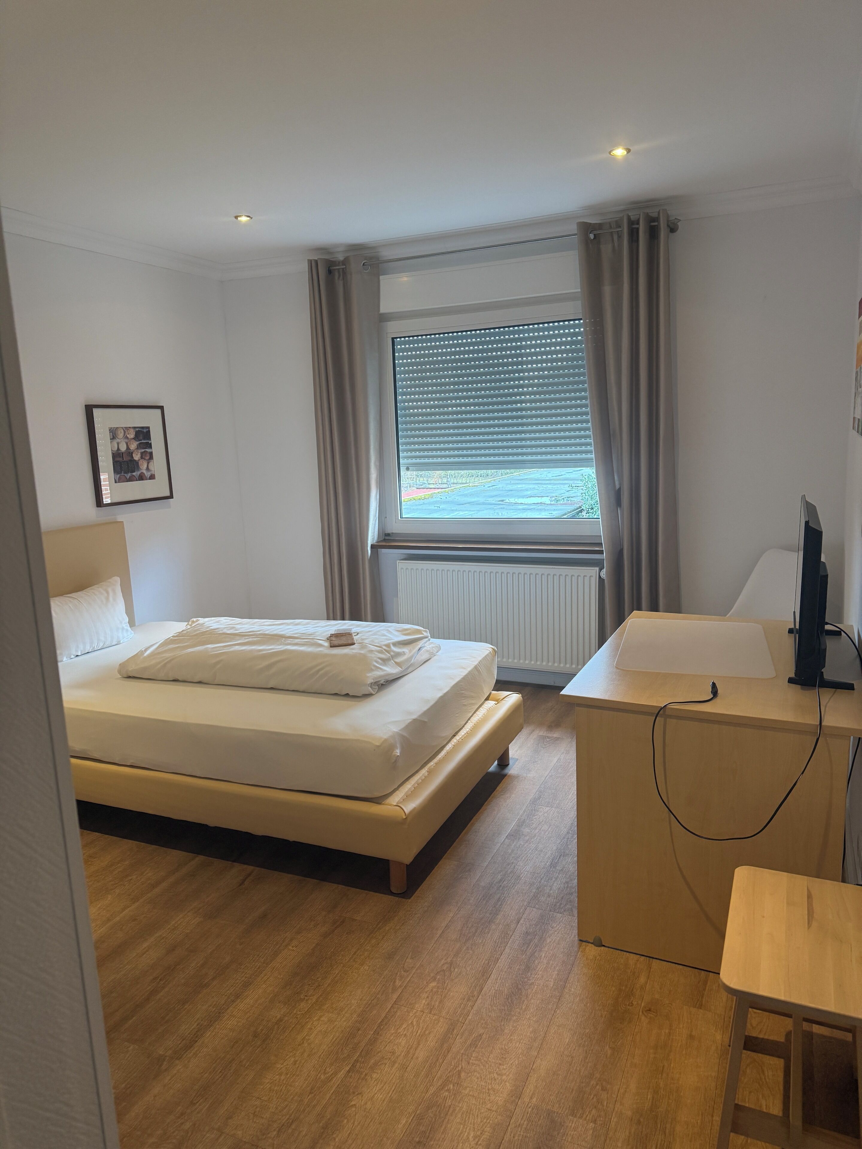 A bedroom with a bed, a desk, a chair, a window with curtains, and a radiator.