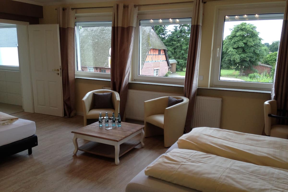 A hotel room with a bed, two armchairs, a small table with bottles, and large windows with curtains.