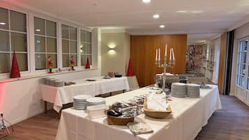 A banquet hall with multiple tables set for dining, a candelabra centerpiece, and a large window with blinds.