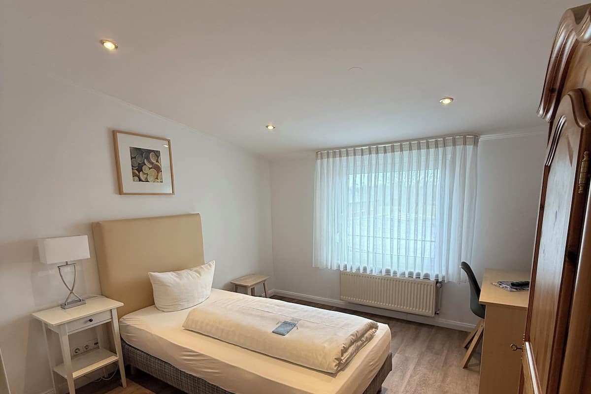 A bedroom with a bed, bedside table, lamp, picture on the wall, window with curtains, radiator, and a desk with chair.