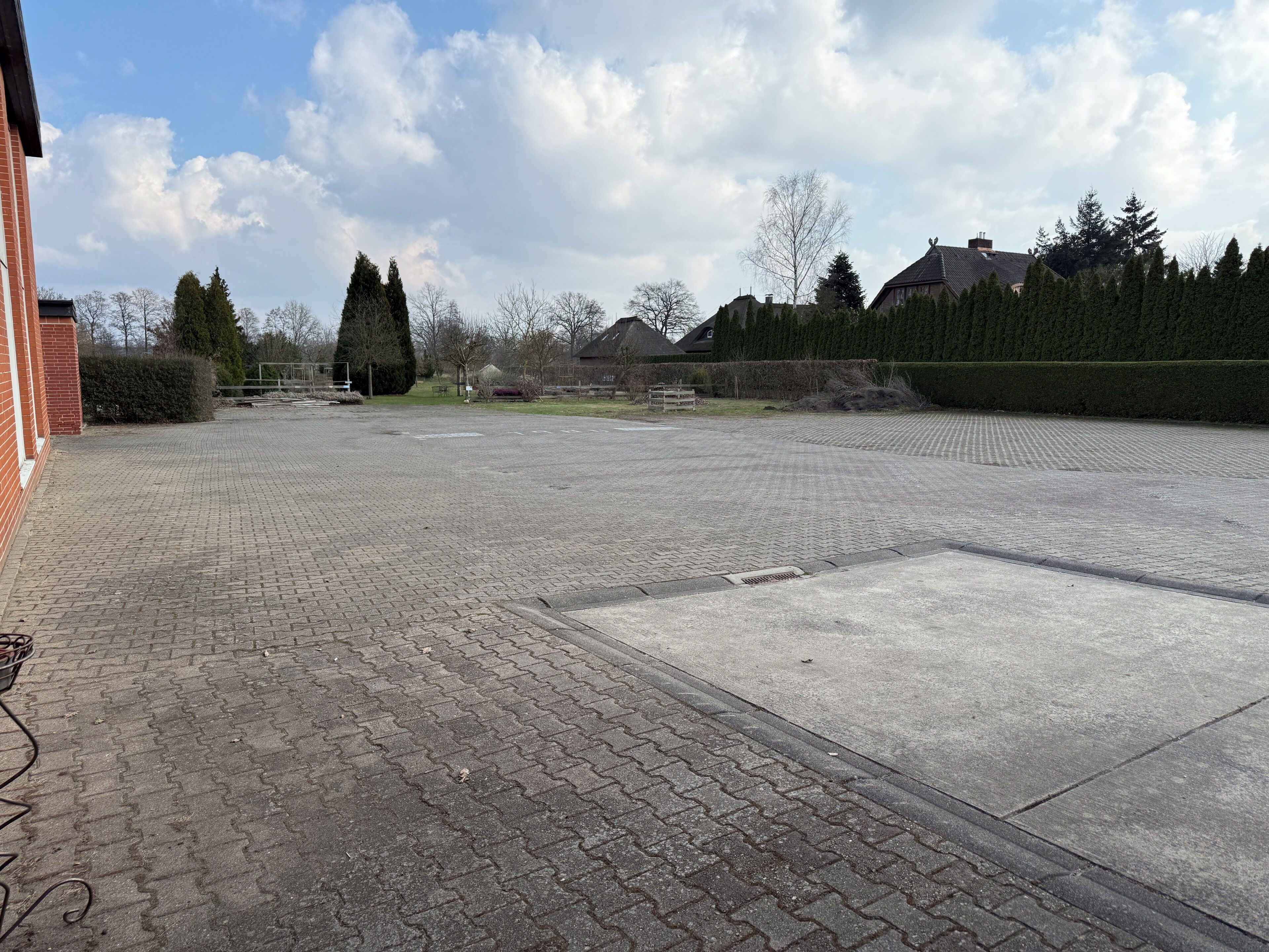 A paved courtyard with a brick wall on one side, a grassy area, and a row of trees and houses in the background.