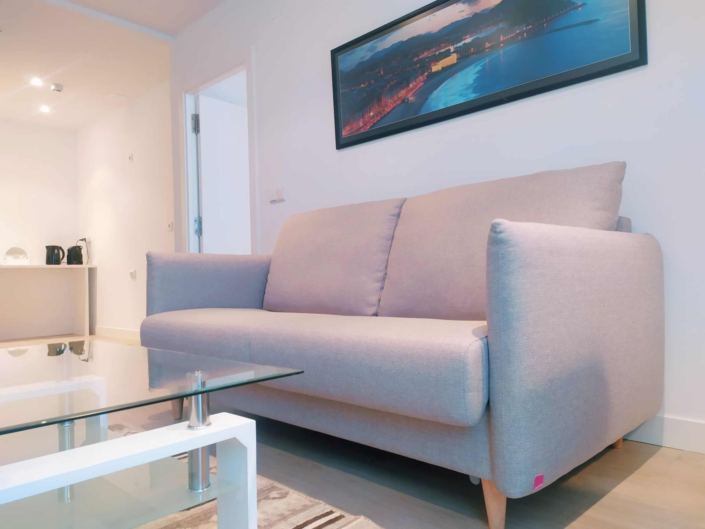 A modern living room with a light grey sofa, a glass coffee table, and a framed cityscape picture on the wall.