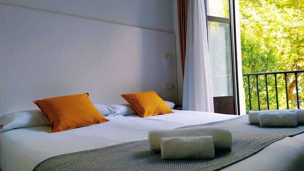 A hotel room with a large bed, two pillows, a blanket, and a view of trees through a window. - Pensión Sarriegi (San Sebastián)