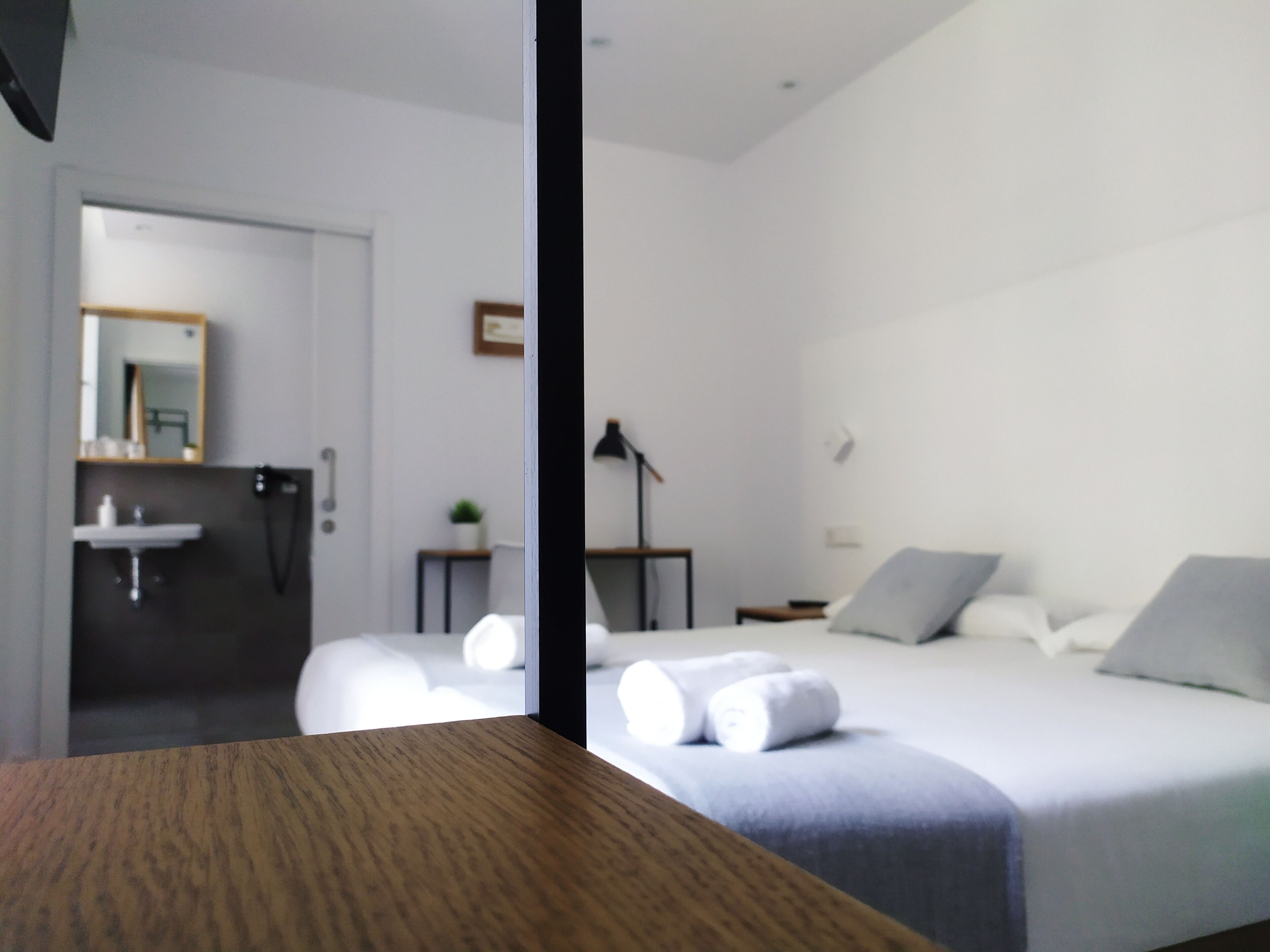Una habitación de hotel moderna con una mesa de madera, una cama con ropa blanca, una mesita de noche, una lámpara y un baño con lavabo y espejo.