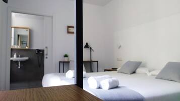 Una habitación de hotel moderna con una mesa de madera, una cama con ropa blanca, una mesita de noche, una lámpara y un baño con lavabo y espejo.
