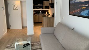 A modern living room with a grey sofa, a glass coffee table, and a remote control on it. A door with the number 12 is visible, leading to a kitchen with wooden cabinets and a black countertop. - Pensión Sarriegi (San Sebastián)