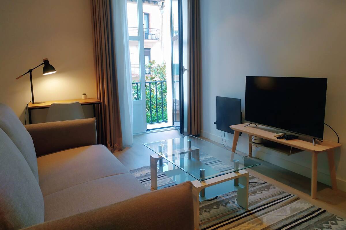 Un salon moderne avec un canapé, une table basse en verre, un téléviseur à écran plat posé sur un meuble, une lampe et une vue sur le balcon extérieur.