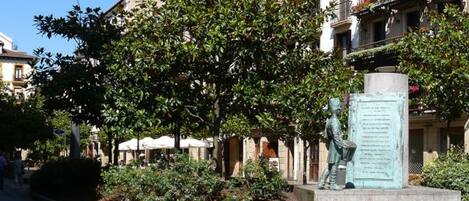 A public square with a statue, surrounded by trees and buildings with balconies.