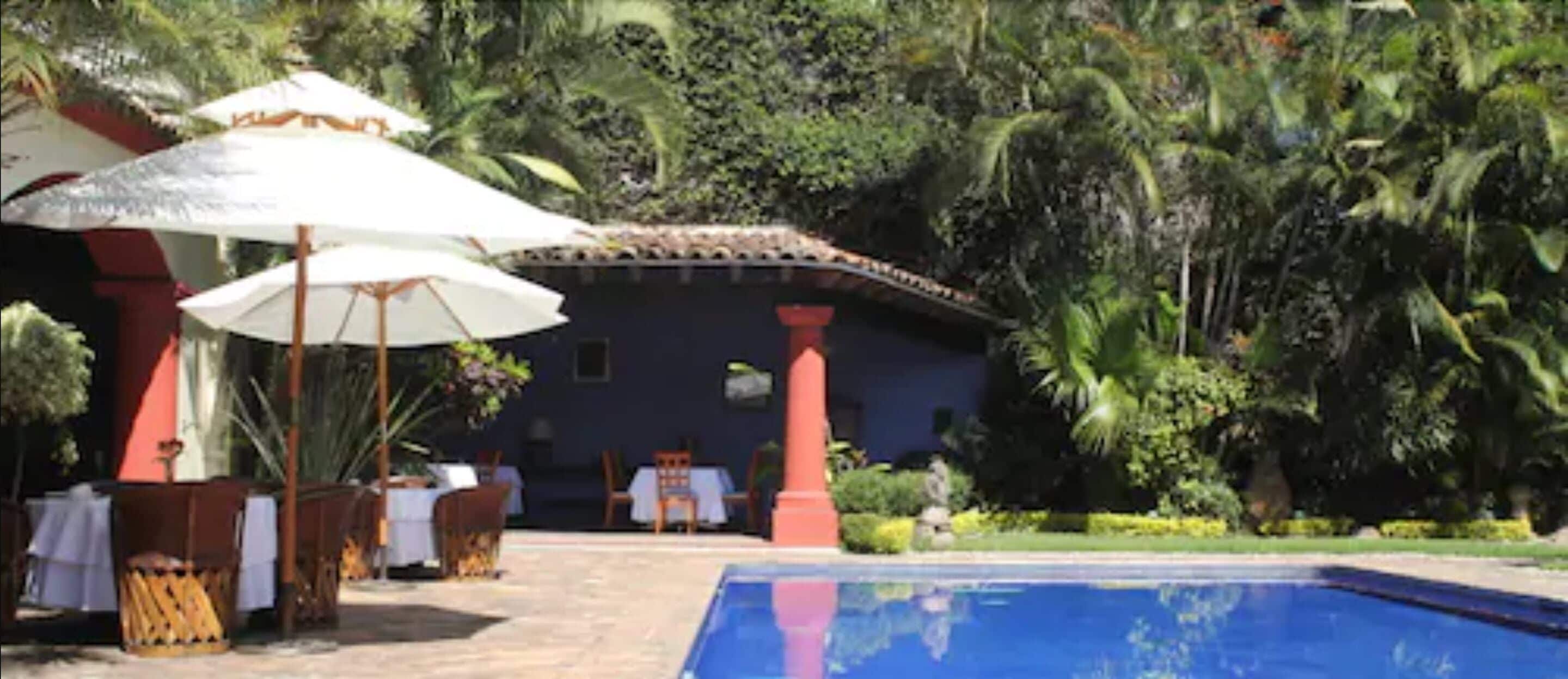 A poolside area with umbrellas, tables, and chairs, surrounded by lush greenery and a building with a tiled roof.